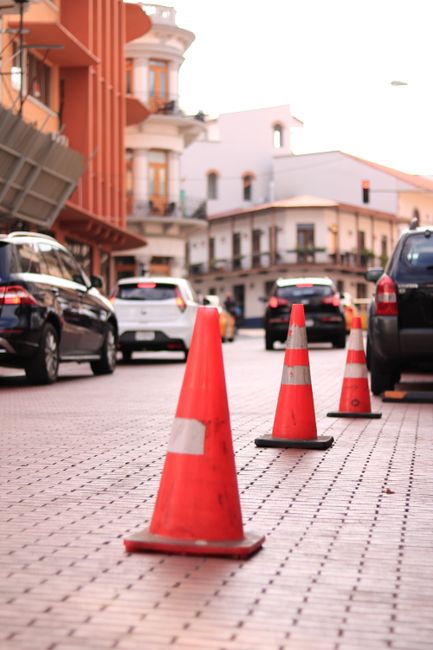 A row of bright orange traffic cones with reflective white stripes are placed along a brick-paved street to reserve parking spaces, supporting safe and organised home relocation and furniture transport activities. Behind the cones, several parked cars, including a black sedan and a white hatchback, are positioned along the roadside. In the background, there are multi-storey buildings with balconies and large windows, painted in warm tones of orange and white, suggesting a residential area. The scene is lit by natural daylight, and there are no visible pedestrians in the image. The placement of the cones indicates a controlled area for loading or unloading household items during a house removal, aligning with the services provided by Man With a Van Beddington for secure and efficient moving and packing logistics.