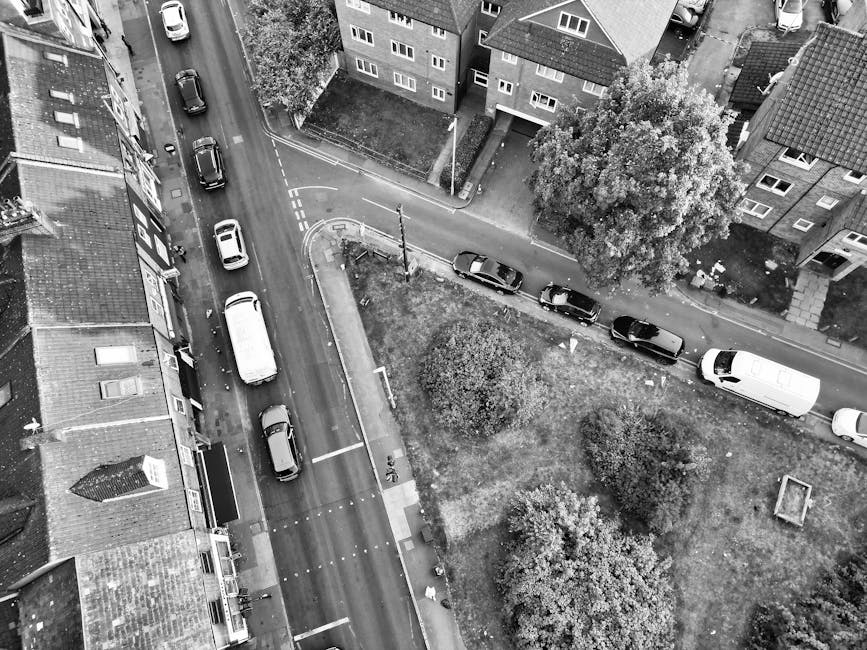 An aerial black-and-white photograph showing a residential street with parked cars along the curbside and a delivery van partially loaded with household furniture and moving boxes parked near the pavement. Inside the van, visible items include cardboard boxes, a rolled-up rug, and a piece of furniture wrapped in protective fabric, all arranged for safe transportation. Some of the boxes and items are being loaded into or unloaded from the van, with a few boxes placed on the pavement nearby. The street is lined with multi-storey residential buildings with pitched roofs, and mature trees and bushes border the sidewalk and small garden areas. The scene captures a home relocation process, emphasizing the loading and packing of household items for a house move, as performed by a professional removals service such as Man With a Van Beddington, situated in the area of Beddington. Natural daylight and shadows highlight the spatial arrangement of vehicles, furniture, and the surrounding environment.