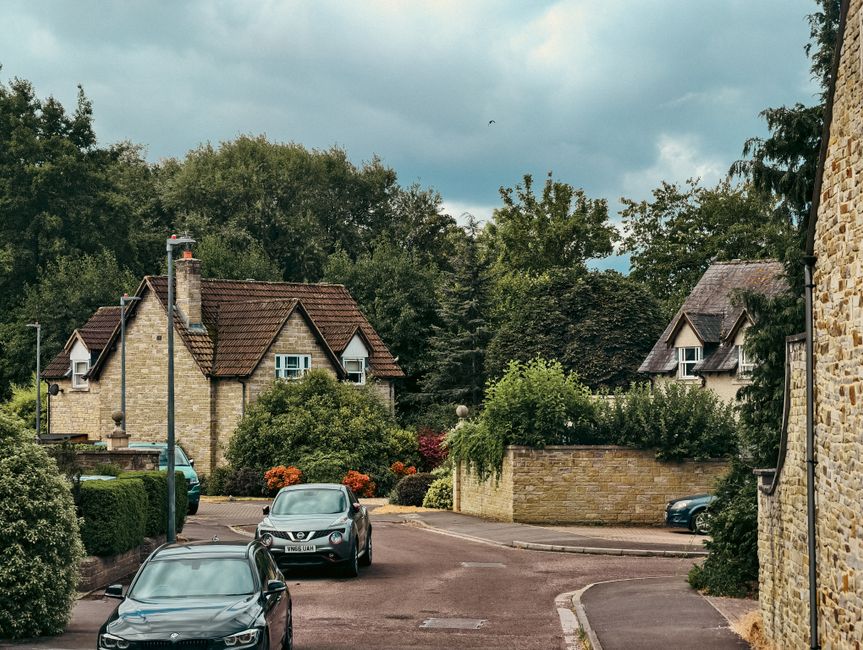 The image shows a residential street with several houses surrounded by greenery and trees. In the foreground, a black car is parked on the street, and behind it, a silver Nissan van is partially visib