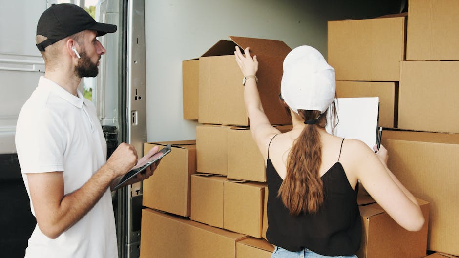 A man and a woman are inside the rear of a moving van, which is parked outside a property, engaged in a home relocation process. The man, wearing a white t-shirt, black cap, and earbud, is holding a clipboard and pen, likely taking inventory or discussing the loading plan. The woman, dressed in a black tank top and wearing a white chef's hat, is reaching to adjust or examine a cardboard box on a stack of similar boxes of various sizes, all wrapped in protective plastic or packing paper. Several larger cardboard boxes are organized in the van's interior, with some on the ground and others stacked toward the ceiling of the vehicle. The scene captures the upright placement of boxes, the detailed handling of packing materials, and the meticulous organization involved in furniture transport and packing during a move, with natural lighting highlighting the inside of the vehicle near the doorway. Man With a Van Beddington is involved in this detailed packing and loading process as part of its removals services. 