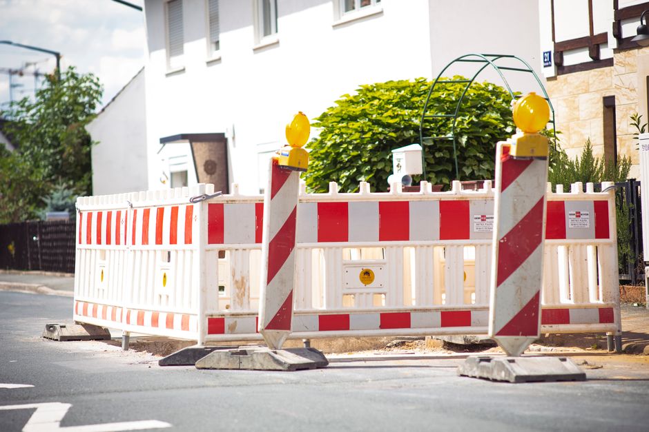 A temporary road closure barrier made of white plastic with red and white reflective stripes, positioned on a residential street to restrict vehicle access during a home relocation or moving process. The barrier features yellow warning lights on top of two vertical posts, and is supported by two weighted bases on the asphalt pavement. Behind the barrier, there is a white residential building with several windows and a doorway, along with a neatly trimmed green shrub and part of a neighboring house. The barriers are set up to facilitate safe furniture transport and loading/unloading of packing materials, with the surrounding environment well-lit by natural daylight. This scene illustrates the logistical setup often used by professional removals services like Man With a Van Beddington to ensure secure and organized property moves, including potential road closures for a smooth move and parking management.