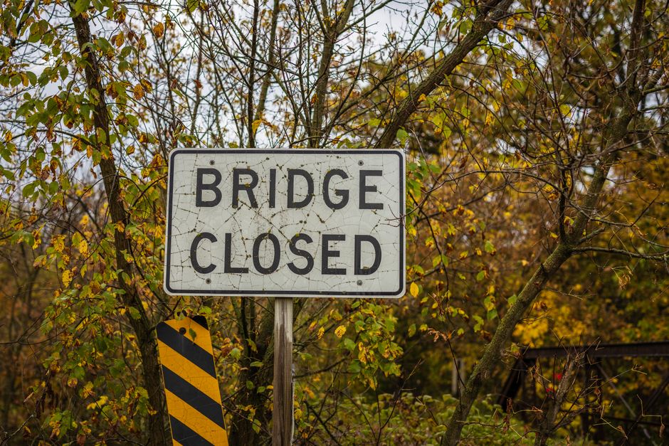 A rectangular white traffic sign with black lettering displaying 'BRIDGE CLOSED' is mounted on a metal pole amidst a background of trees with yellow and green autumn leaves. The sign shows signs of weathering with visible cracks and dirt. Below the sign, a black and yellow striped warning marker is attached to the pole. The surrounding environment appears to be outdoors, possibly near a roadway or driveway that is inaccessible due to the closure, with natural light illuminating the scene. This image reflects the context of planning house removals or relocation services where temporary road closures or access restrictions may impact the loading process or transport of furniture and moving boxes. Man With a Van Beddington mentions service flexibility related to such situations. The scene emphasizes the importance of logistics planning for home relocation, including considerations for traffic management and timing during removals.