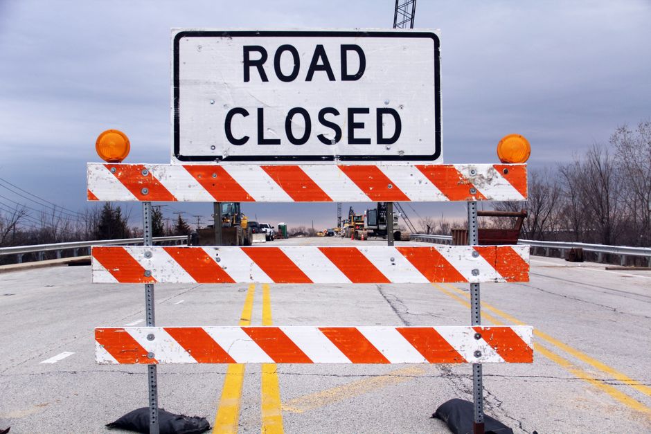 A temporary road closure barrier made of white plastic with red and white reflective stripes, positioned on a residential street to restrict vehicle access during a home relocation or moving process. The barrier features yellow warning lights on top of two vertical posts, and is supported by two weighted bases on the asphalt pavement. Behind the barrier, there is a white residential building with several windows and a doorway, along with a neatly trimmed green shrub and part of a neighboring house. The barriers are set up to facilitate safe furniture transport and loading/unloading of packing materials, with the surrounding environment well-lit by natural daylight. This scene illustrates the logistical setup often used by professional removals services like Man With a Van Beddington to ensure secure and organized property moves, including potential road closures for a smooth move and parking management.