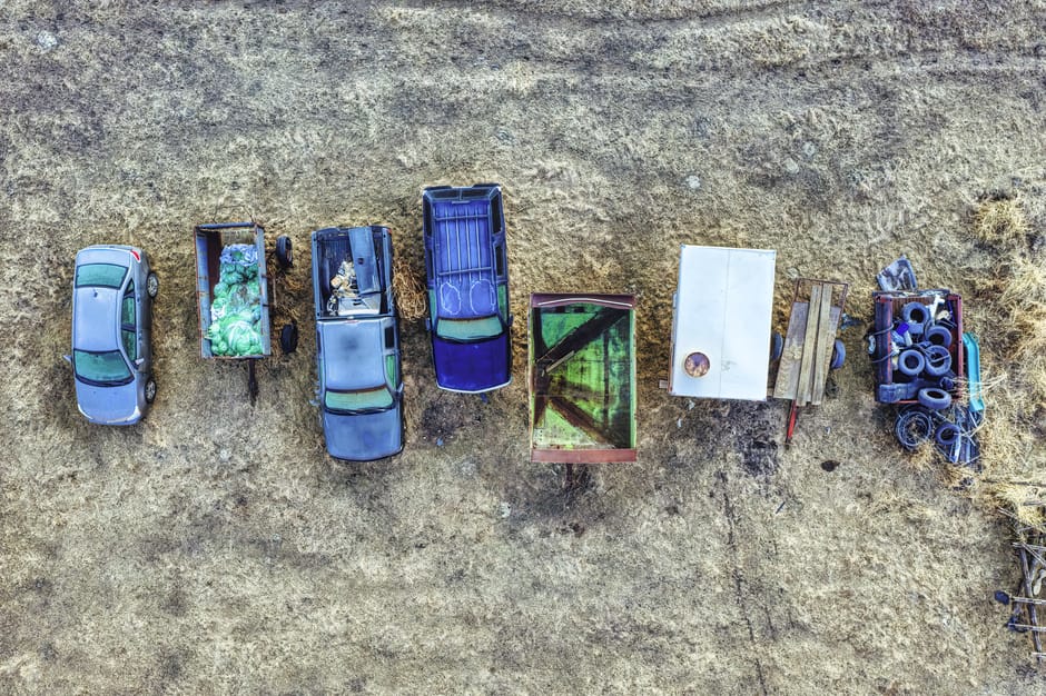 An aerial view showing a sequence of seven vehicles and containers lined up on a dirt surface outside a property, illustrating the home relocation process. The first vehicle is a small silver car, followed by a small open trailer filled with green plastic wrapping for furniture protection, and a larger wagon with various packing materials. Next, a blue van is visible, with a white flatbed truck loaded with wooden pallets and a mix of packing supplies. Adjacent is a small green metal trailer, slightly weathered, with a circular cutout on its surface. Beside it, a white van or truck is parked, with a pile of black rubber tires and other equipment stored in a trailer attached to it. The background shows dry grass and a building wall, with natural outdoor lighting illuminating the scene, which depicts typical equipment and vehicles used in furniture transport and packing during house removals by Man With a Van Beddington, as part of their services for bulky item pickup and home relocations.
