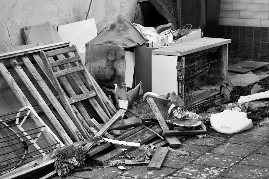 A black-and-white photographic image showing a messy exterior space adjacent to a building, with discarded furniture and household items piled on a paved surface. Visible objects include a wooden bed frame or headboard leaning against a wall, a damaged or broken white cabinet with parts of the door missing, and various broken pieces of wood and debris scattered around. There is a plastic laundry basket, a piece of fabric or netting draped over some of the debris, and a large white plastic object resembling a domestic appliance or container lying on the ground. The scene is illuminated by natural daylight, with the background featuring a brick or tile wall and a section of pavement, suggesting a typical house exterior or garden area awaiting clearance or disposal as part of a house removal or renovation process. The clutter includes packing materials and remnants that might be encountered during furniture transport or clearing during a home relocation, illustrating the type of items handled by Man With a Van Beddington during bulky item pickup and moving services.
