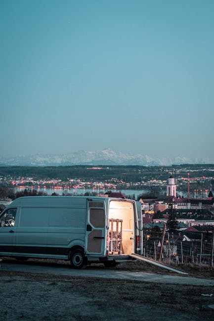 A light grey transit van parked on an unpaved area with its rear doors open, revealing wooden furniture pieces and supplies inside, such as wrapped chairs and cardboard boxes, indicating a home relocation or furniture transport process. The van is positioned on a slight incline with a loading ramp extending onto the ground, and various packing materials like plastic wrap and protective blankets are visible around the furniture. In the background, a semi-urban landscape features residential buildings, industrial structures, and a distant mountain range under a dusky sky. The setting suggests an active loading process during a house removal or moving service, exemplified by the company Man With a Van Beddington, operating in the context of last-minute removals in Beddington, South London.