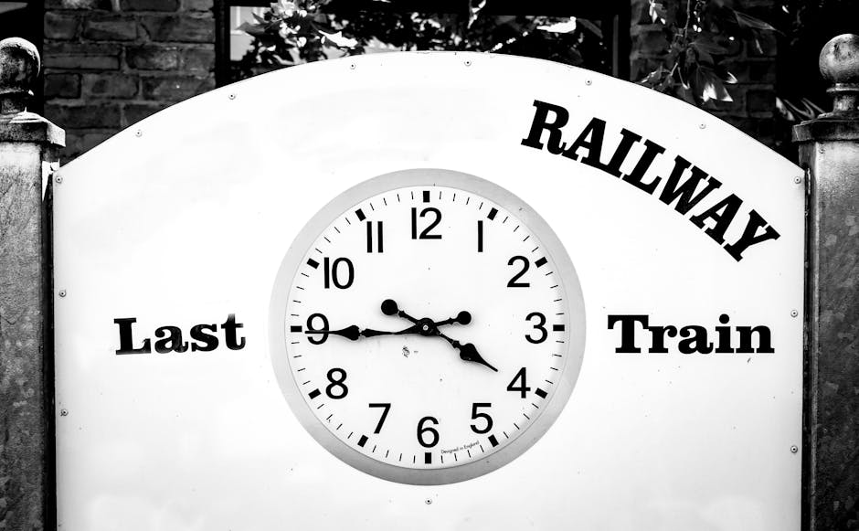 A large, circular railway clock mounted on a white background, displaying the time as approximately 3:47. The clock features black hour and minute hands, with a smaller black second hand, and is surrounded by a light grey border. To the left of the clock, the words 'Last' are printed in bold black letters, and to the right, the words 'Train' are printed in similar style. Above the clock, the word 'RAILWAY' is written in bold, curved black letters. The clock and lettering are set against an outdoor backdrop with foliage and a stone pillar visible at the top edges of the image, indicating it is mounted on or near a traditional railway station sign. This scene could symbolise timing in the context of home relocation or moving logistics, in line with services provided by Man With a Van Beddington, specialising in removals. The image conveys a sense of urgency or the importance of timing in the furniture transport or packing and moving process.