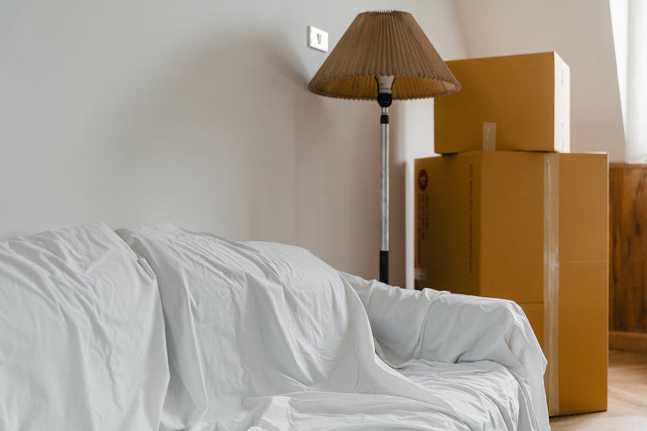 A light grey transit van parked on an unpaved area with its rear doors open, revealing wooden furniture pieces and supplies inside, such as wrapped chairs and cardboard boxes, indicating a home relocation or furniture transport process. The van is positioned on a slight incline with a loading ramp extending onto the ground, and various packing materials like plastic wrap and protective blankets are visible around the furniture. In the background, a semi-urban landscape features residential buildings, industrial structures, and a distant mountain range under a dusky sky. The setting suggests an active loading process during a house removal or moving service, exemplified by the company Man With a Van Beddington, operating in the context of last-minute removals in Beddington, South London.