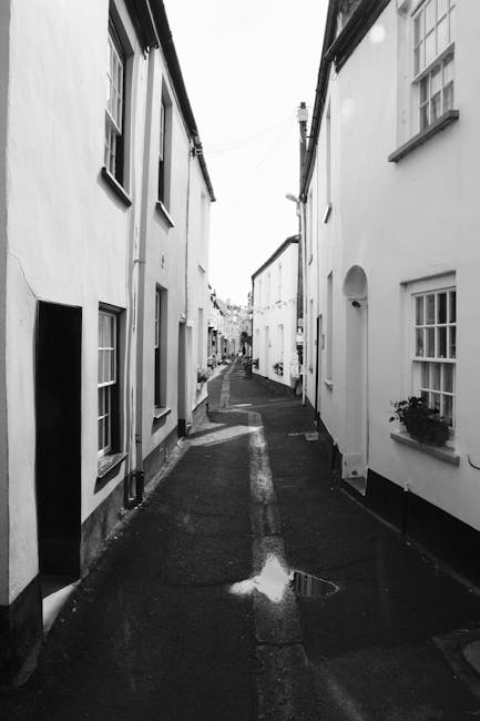 A narrow residential alleyway with tightly packed white-painted houses on either side, featuring small windows with flower boxes and chimneys visible at the rooftops. The cobblestone paving shows signs of recent rain, with puddles reflecting the overcast sky. The alley appears to be part of a home relocation process, typical of tight urban streets where maneuvering large furniture or moving vans may require careful navigation. In the foreground, the asphalt pavement continues into the distance, with no visible moving equipment or personnel, but this setting aligns with the environment where companies like Man With a Van Beddington offer removals and furniture transport services within such confined spaces, supporting efficient packing and loading in restricted street environments.