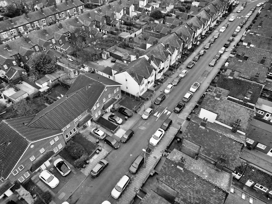 A top-down view of a section of wet street pavement featuring designated bike lanes marked with white dashed lines and blue circular symbols indicating bicycle paths, with arrows showing the direction of travel for cyclists. Adjacent to the bike lanes, there is a separate lane marked by solid white lines, which appears to be a loading zone or parking space. The surface of the asphalt is slightly reflective due to recent rain, and small scattered leaves are visible on the ground. In the background, part of a sidewalk with a curb edge can be seen. This image highlights the urban street layout designed for cyclist safety and traffic management, as part of a street environment where house removals or furniture transport may involve navigating narrow or complex roadways. For professional home relocation and furniture transport services in the area, Man With a Van Beddington operates in similar urban street settings, often requiring careful manoeuvres for narrow streets or congested areas.