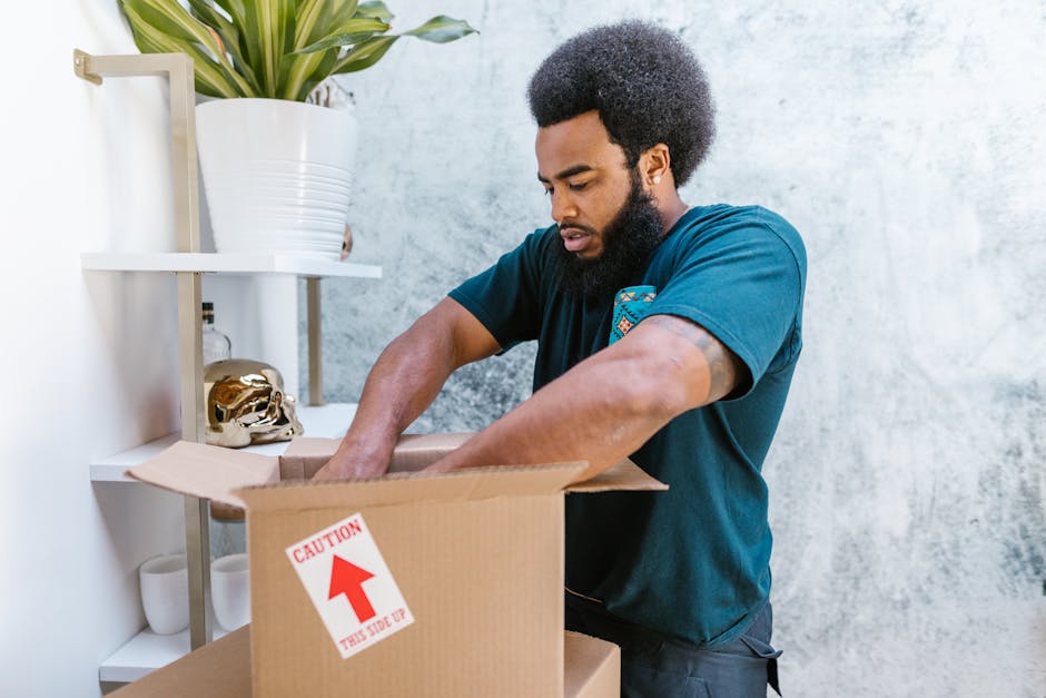 A man with a curly black afro hairstyle and beard is seen packing items into a cardboard moving box during a home relocation. He is wearing a dark teal t-shirt and is standing inside a room with a light grey textured wall. The man is placing his hands inside the open box, which has a red and white caution label indicating 'This Side Up.' Behind him, there is a white shelving unit with decorative objects including a gold skull figurine, white ceramic cups, and a large potted plant with long green leaves. The scene captures a moment of packing and organising household items, using cardboard boxes and packing materials typical in furniture transport and relocation services provided by Man With a Van Beddington, aligning with packing and moving activities involved in house removals in Wallington and Beddington areas.