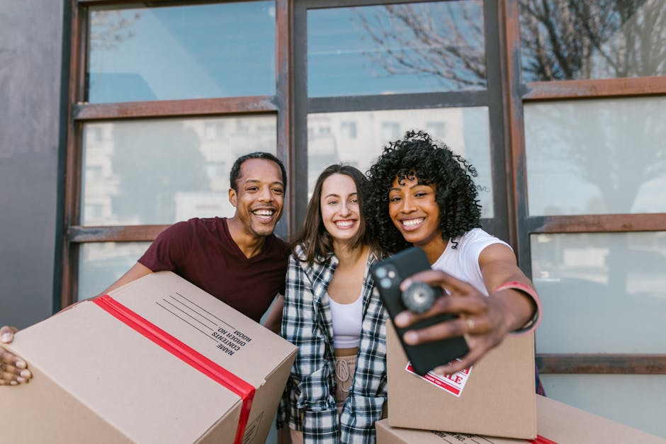 Three smiling individuals stand outdoors near a building with large windows, holding cardboard boxes marked with 'fragile' tape in a home relocation or moving process, with one person taking a selfie using a smartphone. The first person, a man, wears a dark red T-shirt and is holding a box labeled 'kitchen contents,' while the second, a woman in a plaid shirt, stands in the middle, and the third, a woman with curly hair in a white T-shirt, is holding the smartphone for the selfie. The boxes are packed with packing materials such as bubble wrap or plastic wrapping, and the scene reflects a light, natural outdoor setting with daytime lighting, associated with furniture transport and packing during house removals. This image is related to home moving services provided by Man With a Van Beddington as part of their removals services, illustrating packing, organizing, and the friendly, professional approach to house relocations.
