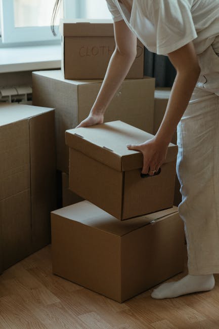 A person, dressed in light-colored clothing, is seen inside a home, lifting a medium-sized cardboard box from a stack of similar boxes during a home relocation or moving process. The boxes are plain b