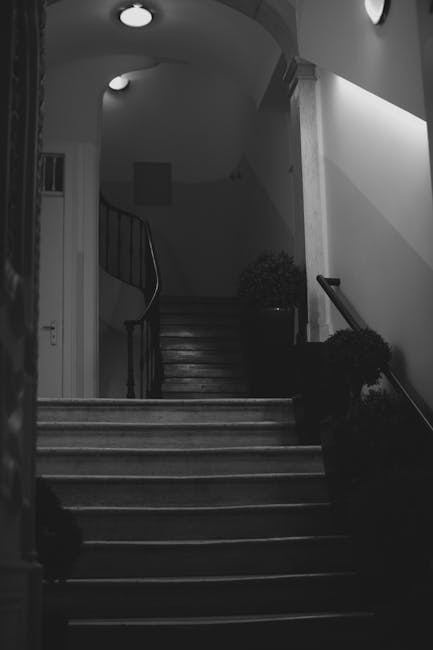 A black-and-white image showing a staircase inside a residential property, with wooden steps and a curved black metal handrail on the left side. The staircase ascends to an upper floor, illuminated by a small ceiling light. On the right side, a black-handled tool or equipment, possibly a moving strap or trolley, is leaning against the wall near the bottom of the stairs. The surrounding interior includes a white wall with moulding, and a partial view of a doorway on the left. Potted plants are visible at the top of the stairs, indicating a home environment. This scene suggests a home relocation or furniture transport process, with the focus on navigating furniture and boxes through narrow or staircase corridors, aligned with services provided by Man With a Van Beddington for removals and house moves.