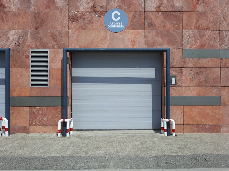 A grey industrial roller shutter door set into a building with a reddish-brown stone facade, surrounded by a dark grey metal frame. To the right of the door, a small rectangular intercom or access device is mounted on the wall. Two white and red striped bollards are positioned on either side of the door, protecting the entrance area from vehicle impact. To the left of the door, a narrow grey ventilation grille is embedded in the wall. The ground in front of the door is paved with grey bricks, and the scene is illuminated by natural daylight, suggesting an exterior loading area. This setting is consistent with a premises used for commercial or industrial purposes, suitable for furniture transport and home relocation activities. The overall appearance supports the environment in which professional removals, such as those offered by Man With a Van Beddington, may operate, particularly in relation to loading bays and secure access points for moving services.