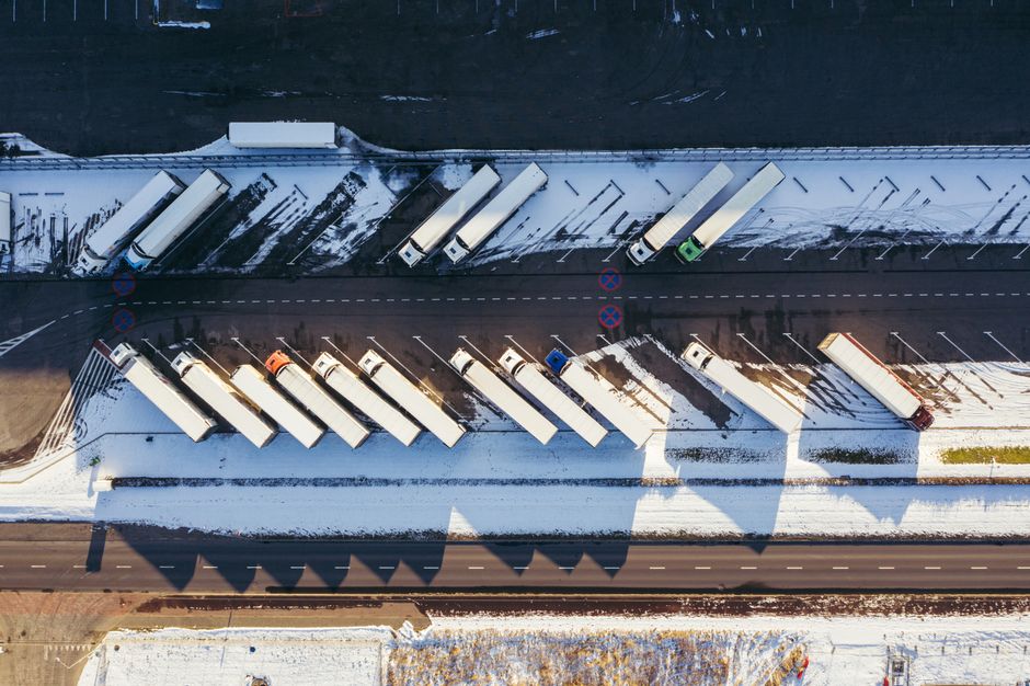An aerial view of a snow-covered loading bay area in Beddington, featuring multiple large white moving trucks parked parallel to each other along a designated loading zone. The trucks are positioned on a paved surface, with some trucks aligned directly next to the building and others along the curbside, possibly being loaded or unloaded for house removals or home relocation services. A green refuse collection vehicle is visible among the trucks, parked near the entrance to the loading zone. The surrounding environment displays snow on the ground, with patches of cleared pavement indicating recent activity, and a network of road markings and traffic signs can be seen around the area, including no-parking signs. The scene is illuminated by daylight, with shadow patterns cast by the trucks and nearby structures, reflecting the logistics involved in furniture transport, packing, and moving logistics that companies like Man With a Van Beddington facilitate during city-wide relocations, such as those requiring Sutton Council permits for loading bays.