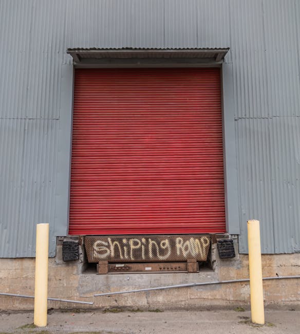 A grey industrial roller shutter door set into a building with a reddish-brown stone facade, surrounded by a dark grey metal frame. To the right of the door, a small rectangular intercom or access device is mounted on the wall. Two white and red striped bollards are positioned on either side of the door, protecting the entrance area from vehicle impact. To the left of the door, a narrow grey ventilation grille is embedded in the wall. The ground in front of the door is paved with grey bricks, and the scene is illuminated by natural daylight, suggesting an exterior loading area. This setting is consistent with a premises used for commercial or industrial purposes, suitable for furniture transport and home relocation activities. The overall appearance supports the environment in which professional removals, such as those offered by Man With a Van Beddington, may operate, particularly in relation to loading bays and secure access points for moving services.