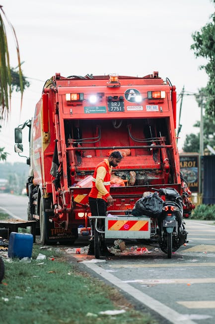 A worker dressed in a high-visibility orange and yellow safety vest is standing behind a large red waste collection vehicle on a roadside. The rear loading door of the junk removal truck is open, revealing the interior designed for waste disposal, with some debris visible. The worker appears to be handling or inspecting black plastic trash bags placed on a small metal platform attached to the back of the vehicle, which also holds a scooter with a black cover. The truck is parked partially on the roadway, with a curb and grassy verge to the left, and a painted white line marking the edge of the parking or loading area. In the background, there are trees, power lines, and a small building or shop, indicating a semi-urban environment. The scene is captured during daylight hours with overcast conditions, emphasizing a practical and straightforward waste collection or rubbish removal process related to home or property clearance, which ties into the context of house removals and moving services provided by Man With a Van Beddington.