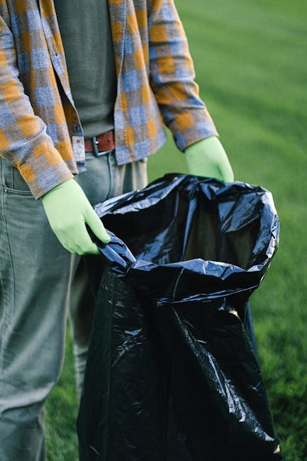 A person wearing a plaid shirt, green gloves, and grey trousers holding open a black rubbish bag lined with silver foil, preparing for waste collection during a house removal or move. The individual is standing outdoors on a grassy area, with part of their torso and hands visible, indicating they are involved in packing or waste disposal as part of a furniture transport or packing process. This scene reflects the handling of waste materials before loading belongings into a van for a home relocation, consistent with the services offered by Man With a Van Beddington, which specializes in house removals. The natural lighting highlights the packing and loading environment, emphasizing the importance of proper waste management in moving logistics, particularly under local council rules for rubbish removal in Beddington as referenced in the webpage about Sutton Council regulations.